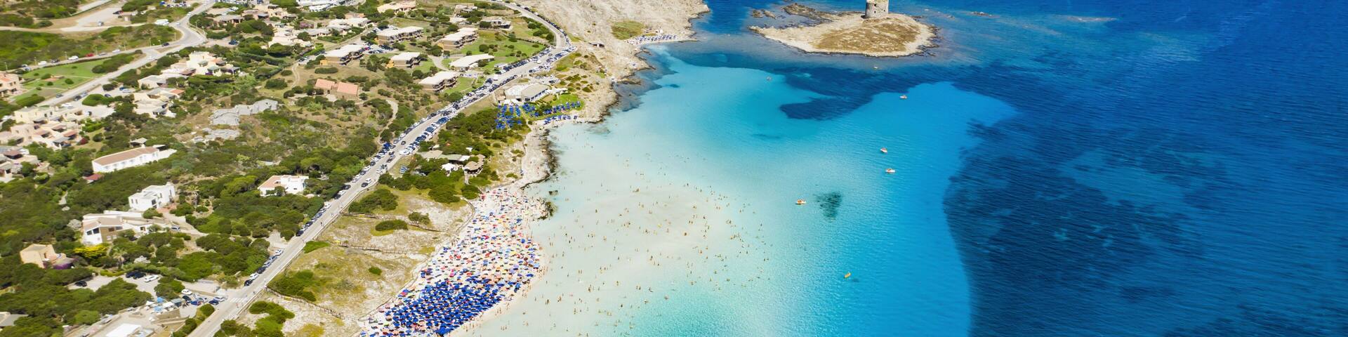 An aerial view of a coastal town with a beach, clear blue waters, and a small island with a tower.