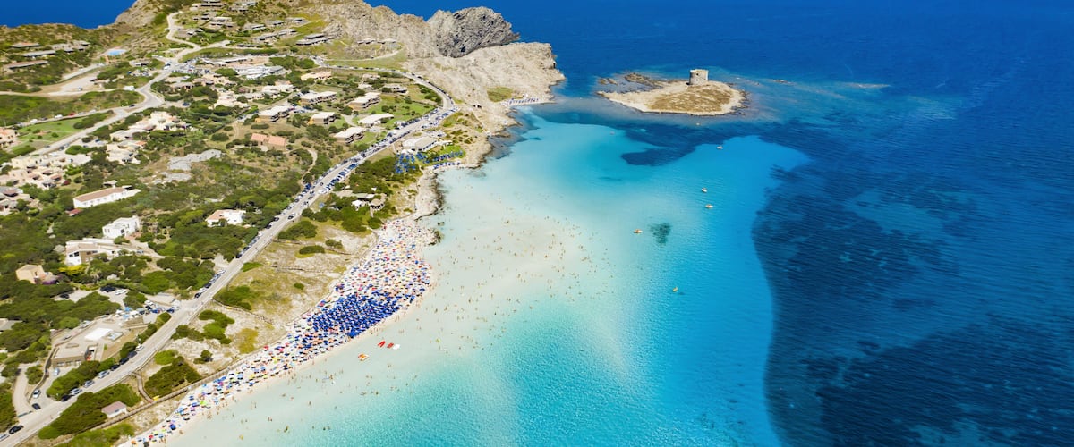 An aerial view of a coastal town with a beach, clear blue waters, and a small island with a tower.