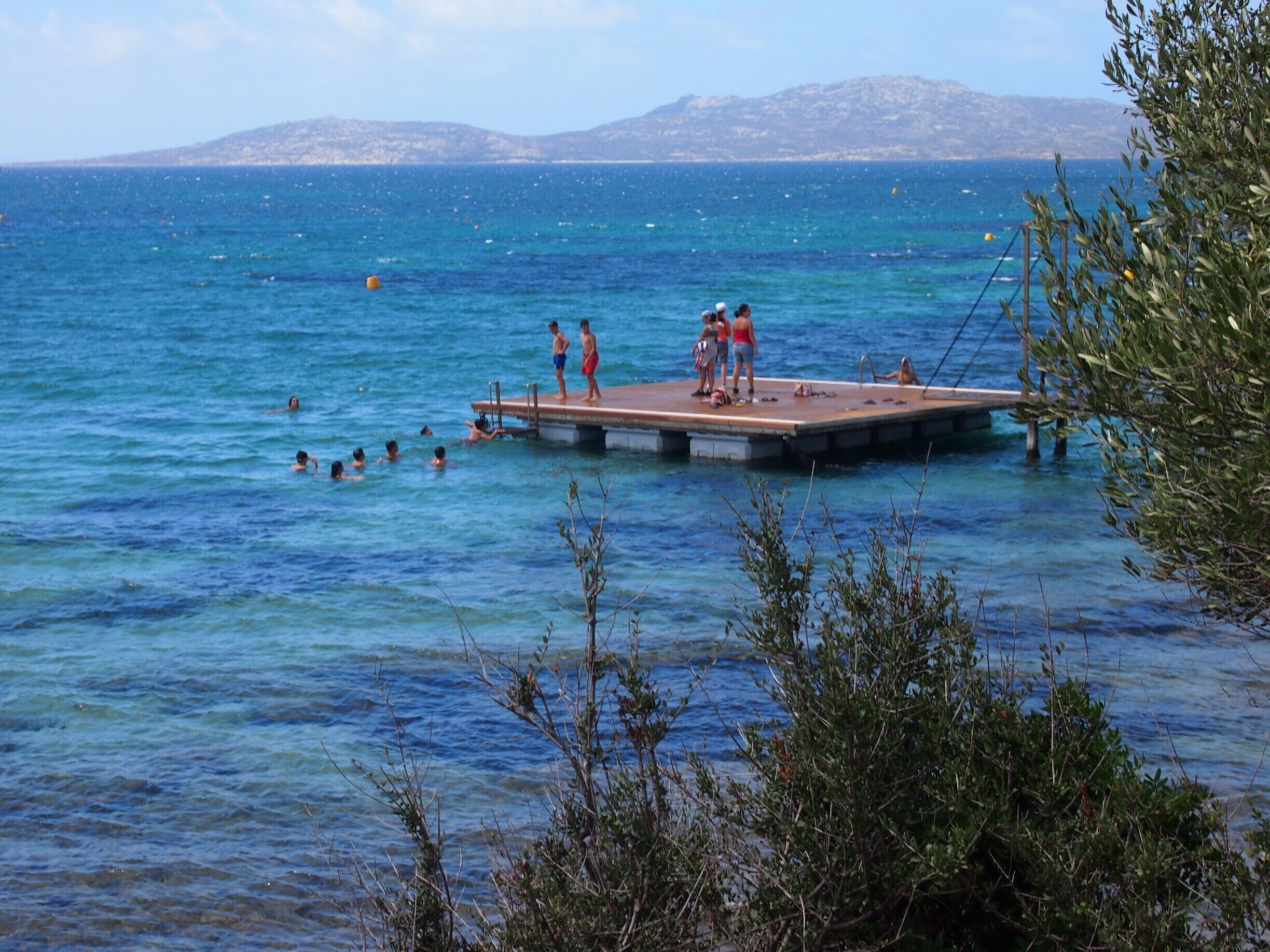 A pier extending into a clear blue sea with people swimming and standing on it.