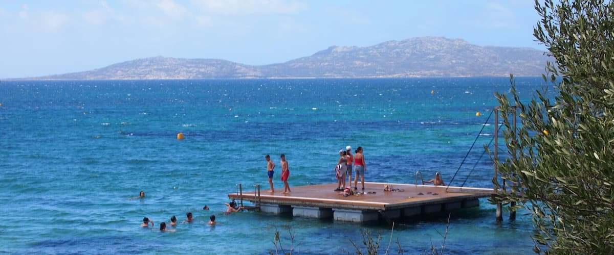 A pier extending into a clear blue sea with people swimming and standing on it.