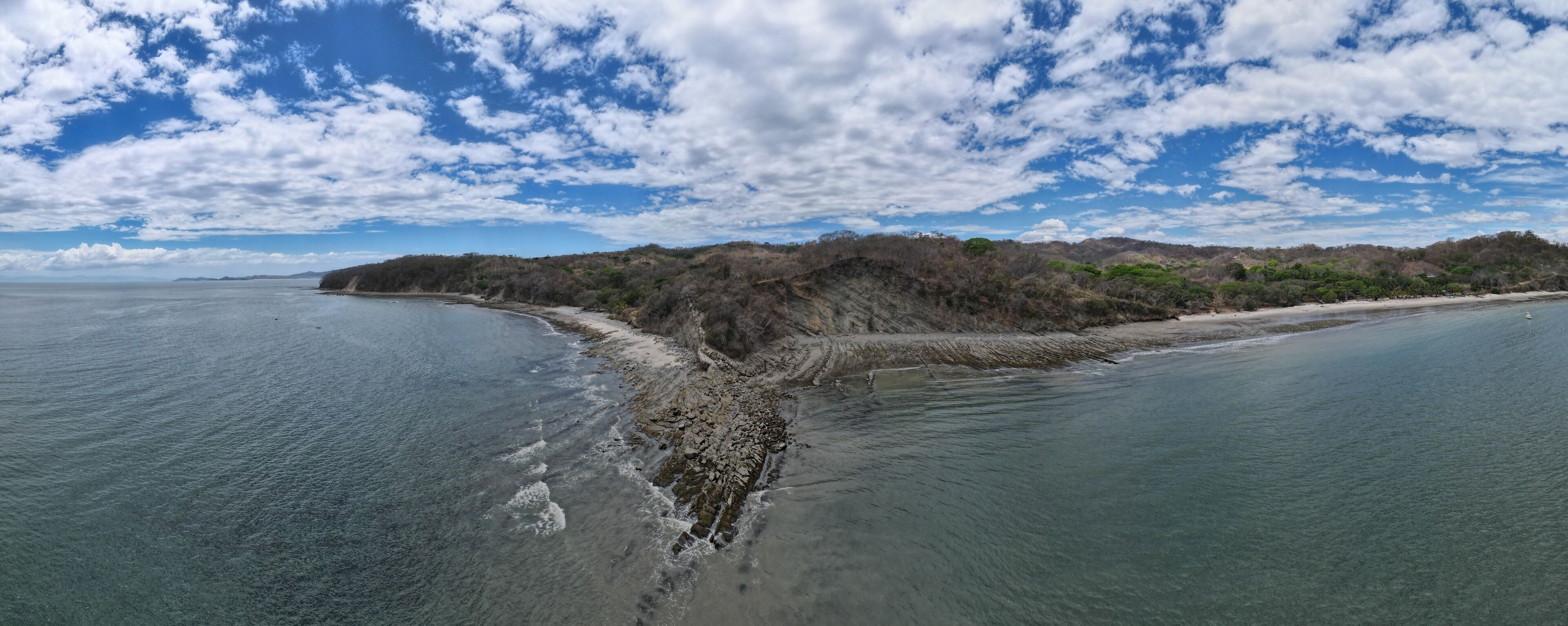A coastal landscape with a sandy beach, rocky shoreline, and a forested hill in the background.