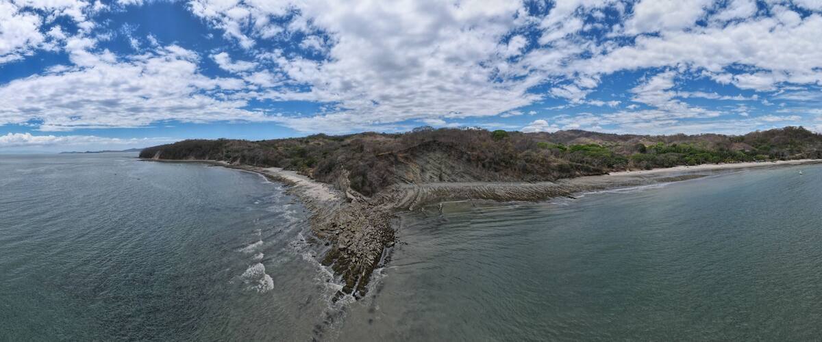 A coastal landscape with a sandy beach, rocky shoreline, and a forested hill in the background.