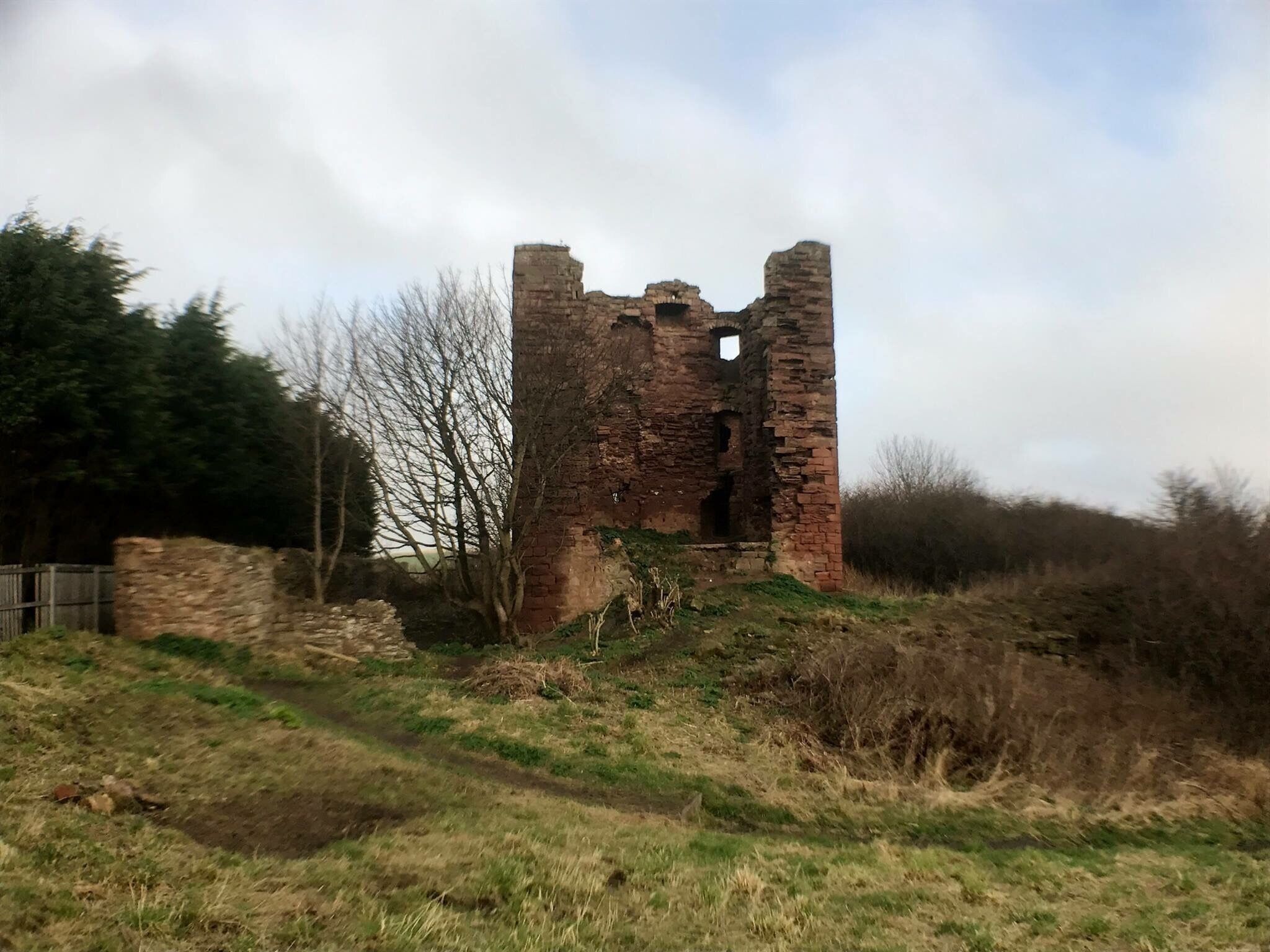 A ruined brick tower amidst a grassy field with trees and a cloudy sky.