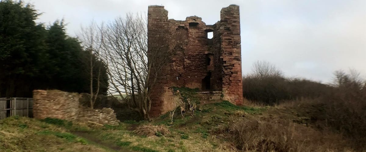 A ruined brick tower amidst a grassy field with trees and a cloudy sky.