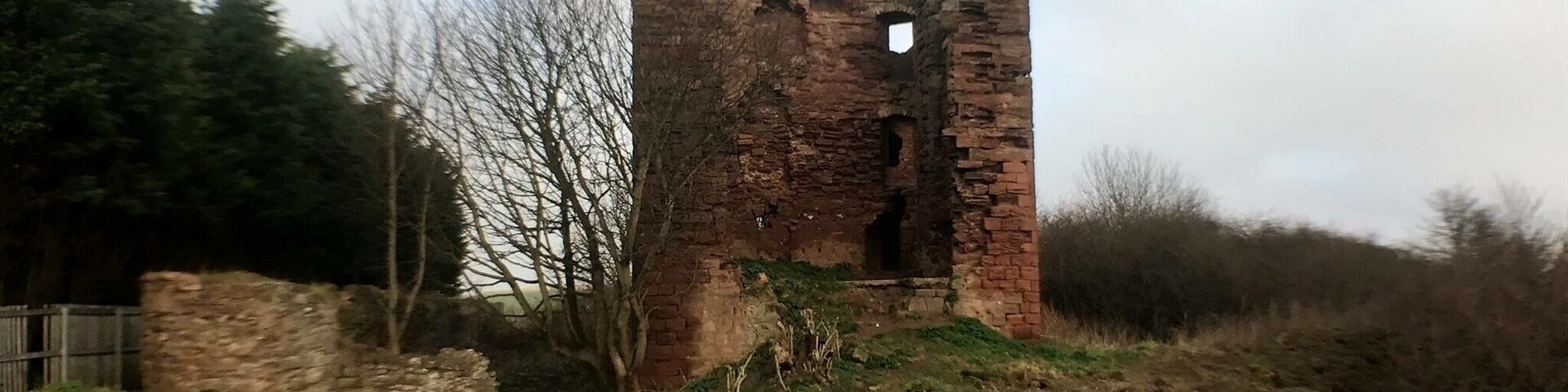 A ruined brick tower amidst a grassy field with trees and a cloudy sky.