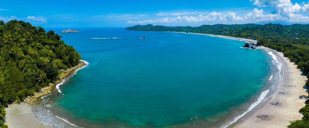 A coastal landscape with a sandy beach, turquoise waters, and dense green forest.