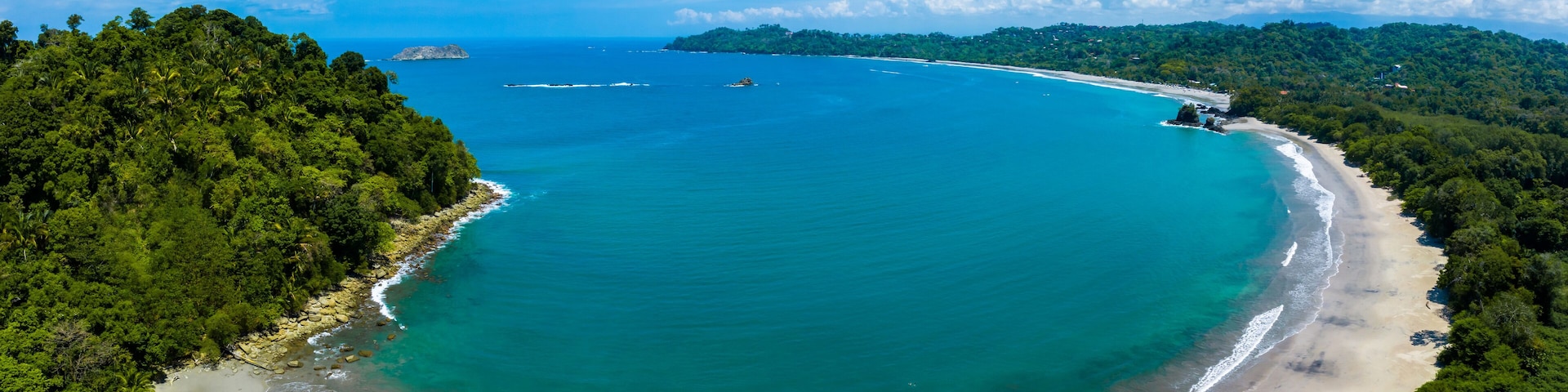 A coastal landscape with a sandy beach, turquoise waters, and dense green forest.