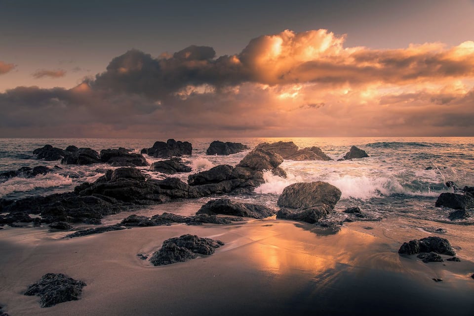 A rocky beach with waves crashing against the shore under a dramatic sunset sky.