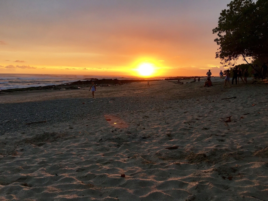 Ein Strand bei Sonnenuntergang, die Sonne tief am Horizont, Menschen, die am Ufer entlanggehen, und eine Felsformation im Hintergrund.