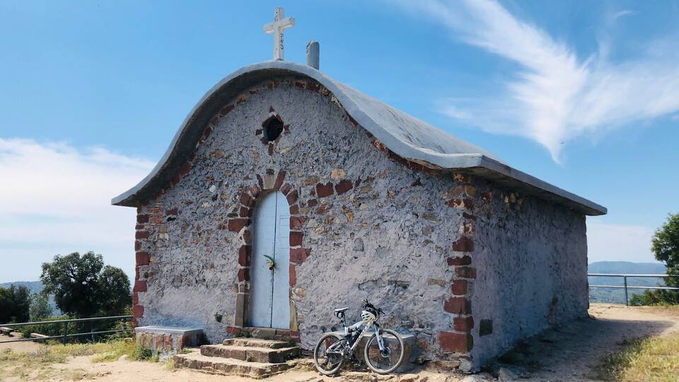 Una pequeña iglesia desgastada por el tiempo con una cruz en la parte superior, una bicicleta estacionada al frente y un cielo azul despejado al fondo.
