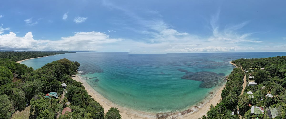 A coastal landscape with a sandy beach, clear turquoise waters, and dense green forest.