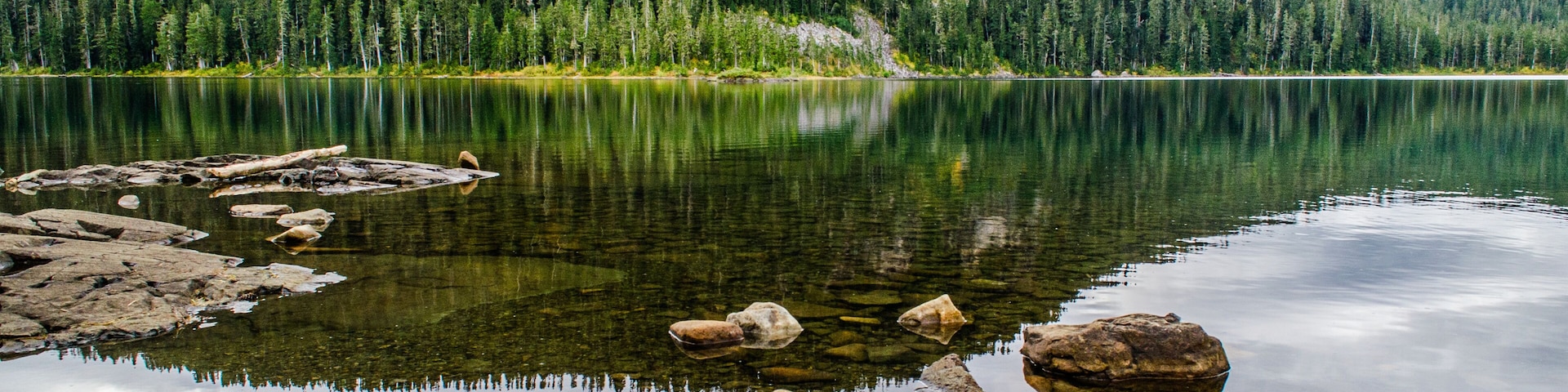 A calm lake with clear reflections, surrounded by dense forest and rocky shores.