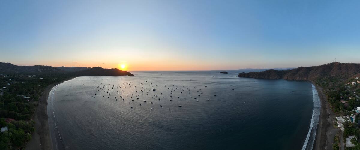 A coastal landscape with a sunset, calm sea, and scattered boats.