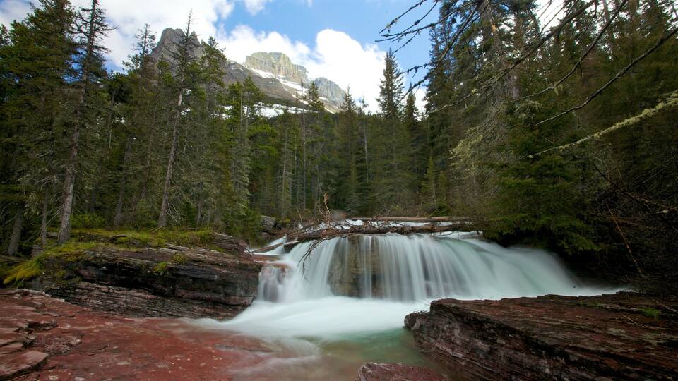 A waterfall cascading over red rocks surrounded by dense forest and mountains in the background.