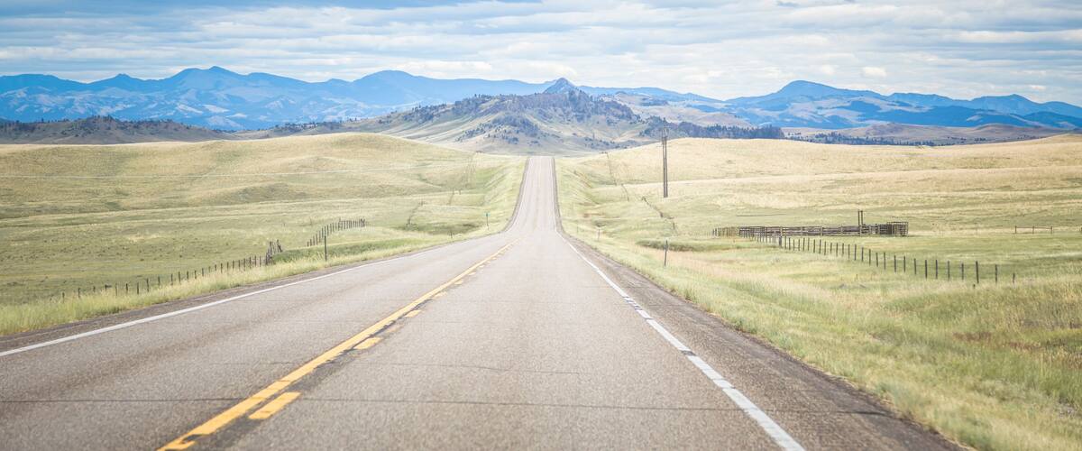 A straight road with yellow center lines, flanked by grassy fields and mountains in the distance under a cloudy sky.