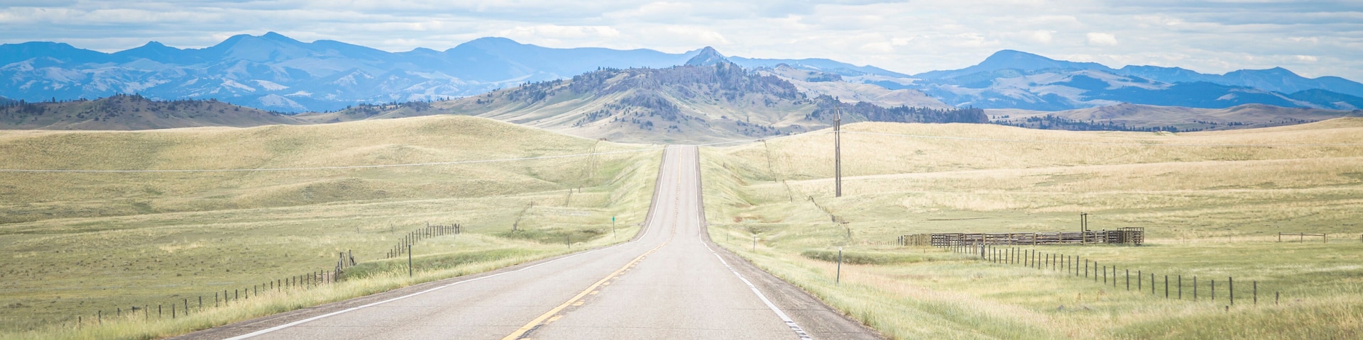 A straight road with yellow center lines, flanked by grassy fields and mountains in the distance under a cloudy sky.