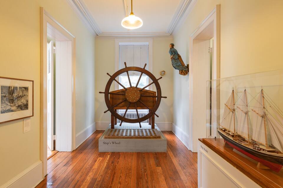A hallway with a ship's wheel, framed maritime paintings, and a ship model.