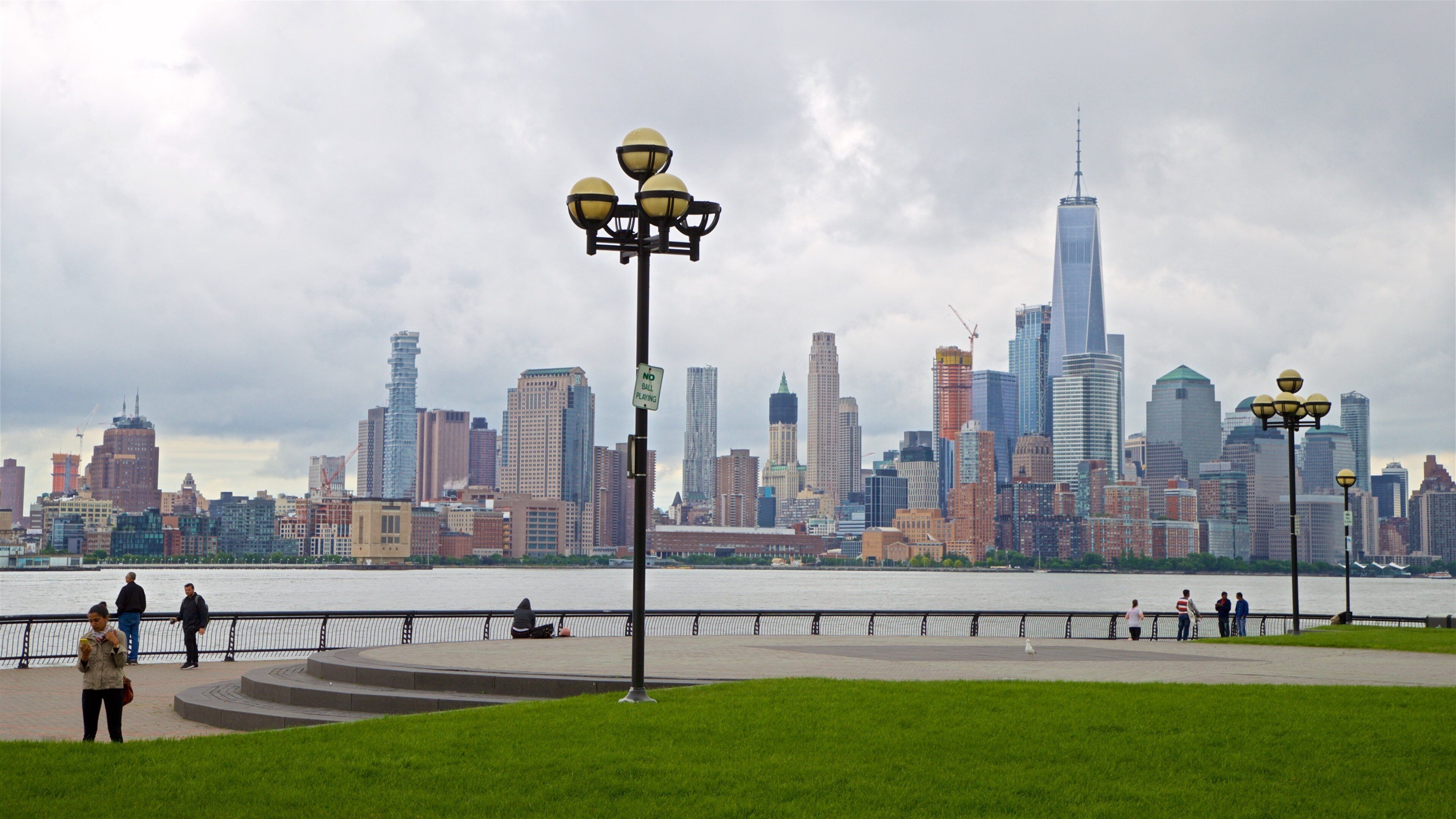 A city skyline with a river in the foreground and a park with people and street lamps.