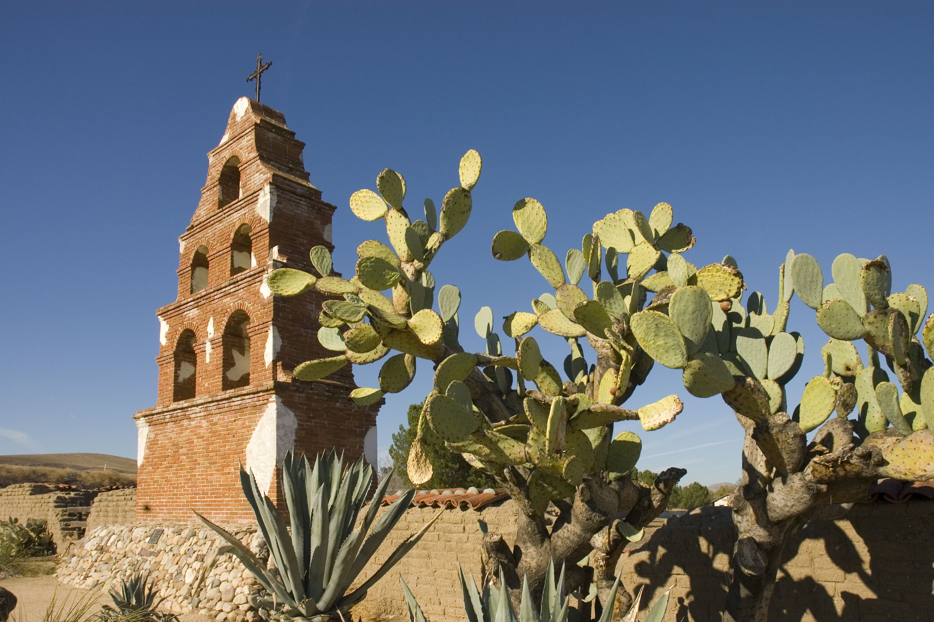 A brick bell tower with a cross, surrounded by cacti and agave plants.