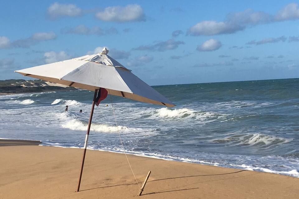 Uma praia com ondas, um guarda-sol branco e céu claro.