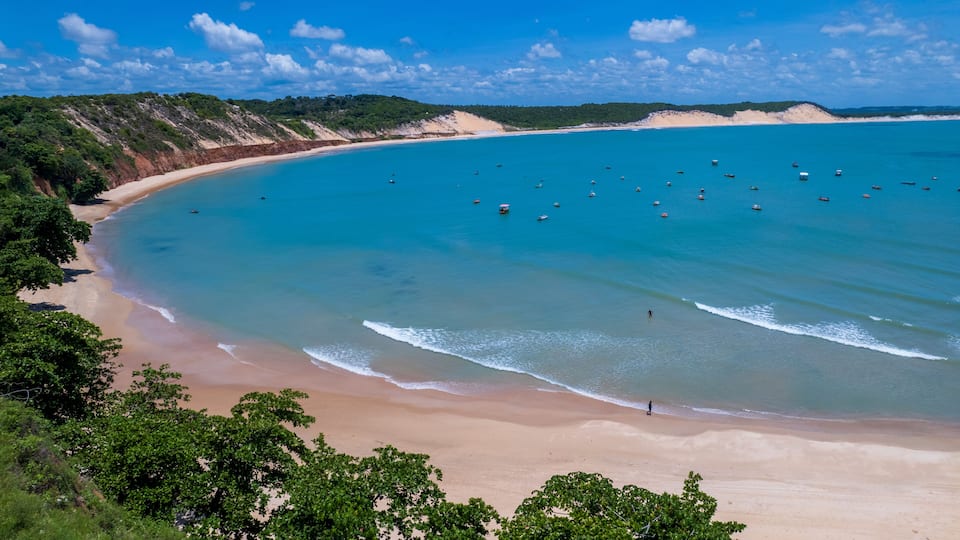 A coastal landscape with a sandy beach, clear turquoise waters, and a hilly backdrop.