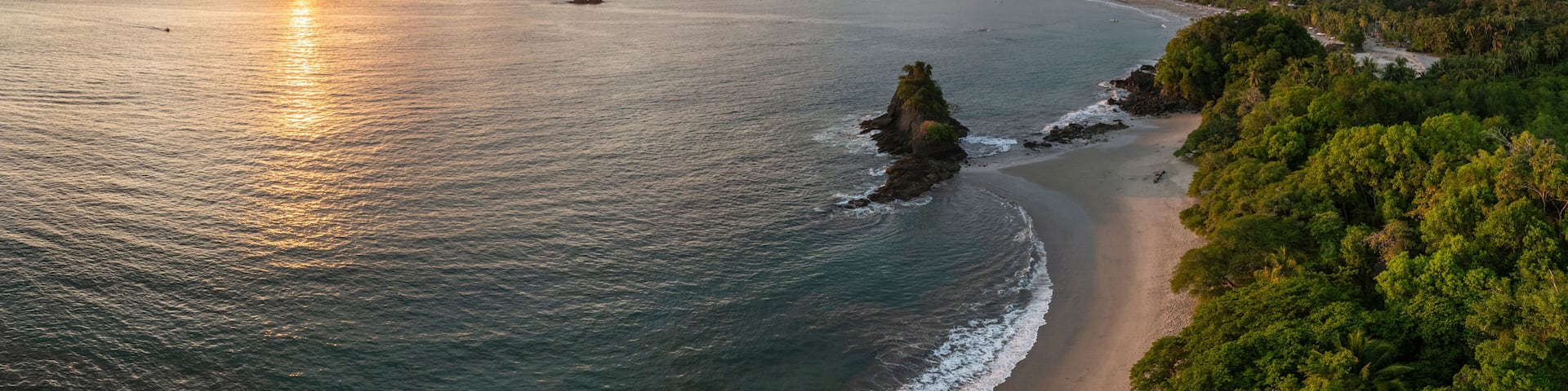 A coastal landscape with a beach, ocean, and forested hills during sunset.