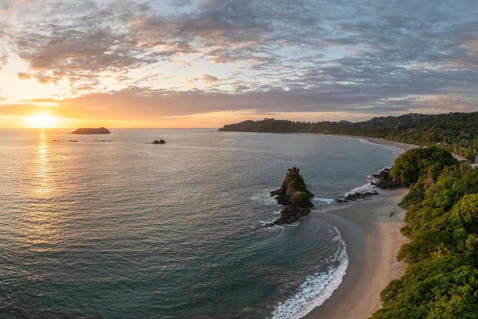 A coastal landscape with a beach, ocean, and forested hills during sunset.