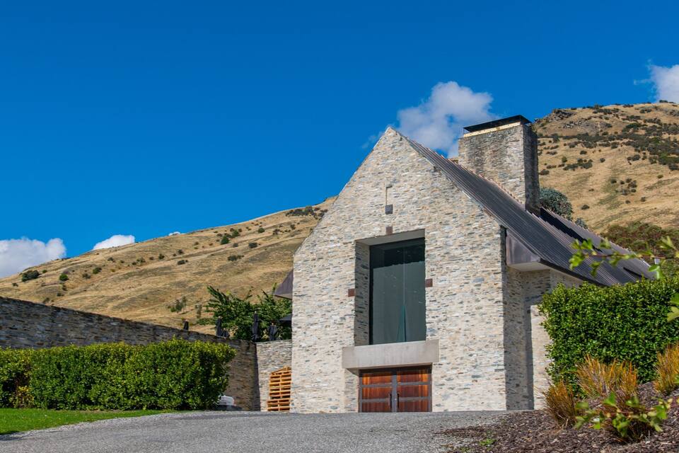 A stone house with a steep roof and a large window, surrounded by greenery and a hill in the background.