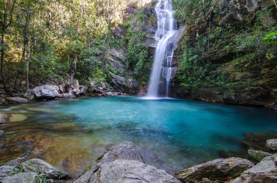 A waterfall cascading into a clear turquoise pool surrounded by rocky terrain and dense green foliage.