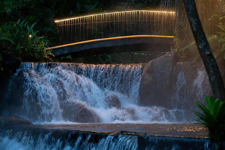 A waterfall cascading over rocks, a bridge with lights, and lush greenery.