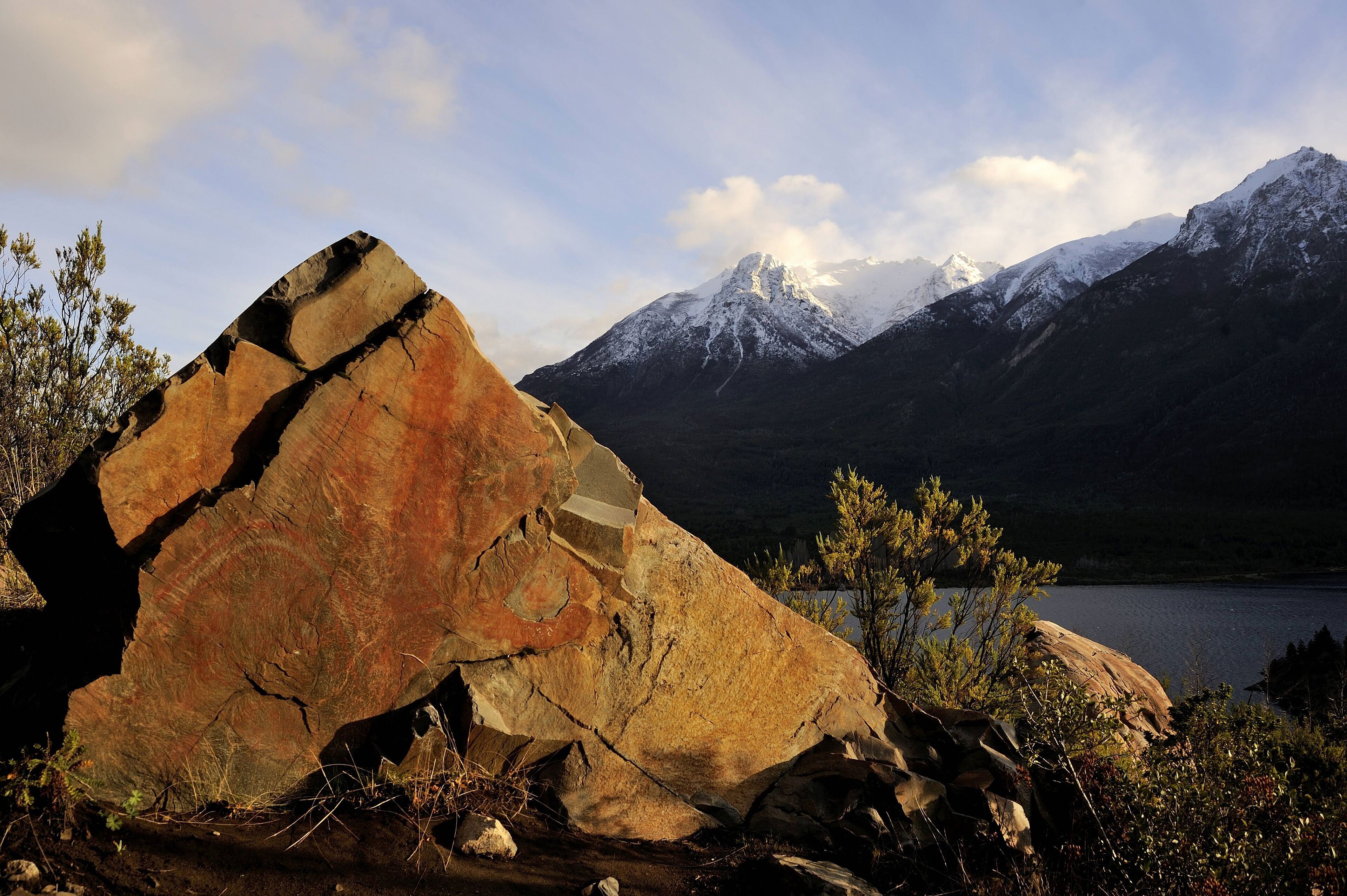 A large rock formation in a mountainous landscape with a lake and snow-capped peaks in the background.