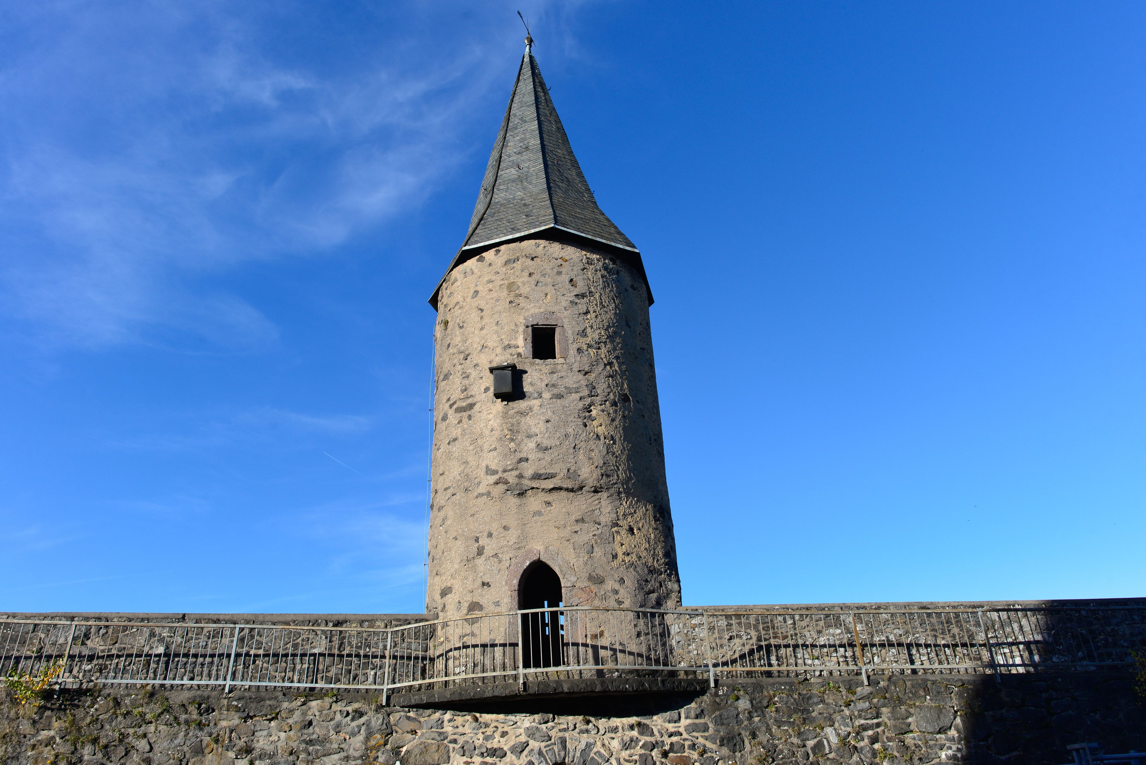 Ein hoher, zylindrischer SteinTurm mit einem spitzen Dach, einem kleinen Fenster und einer Tür, umgeben von einer Steinmauer und einem MetallGeländer.