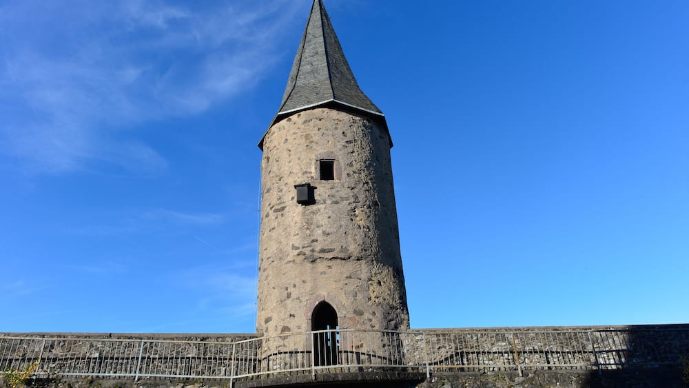 A tall, cylindrical stone tower with a conical roof, a small window, and a doorway, surrounded by a stone wall and a metal railing.