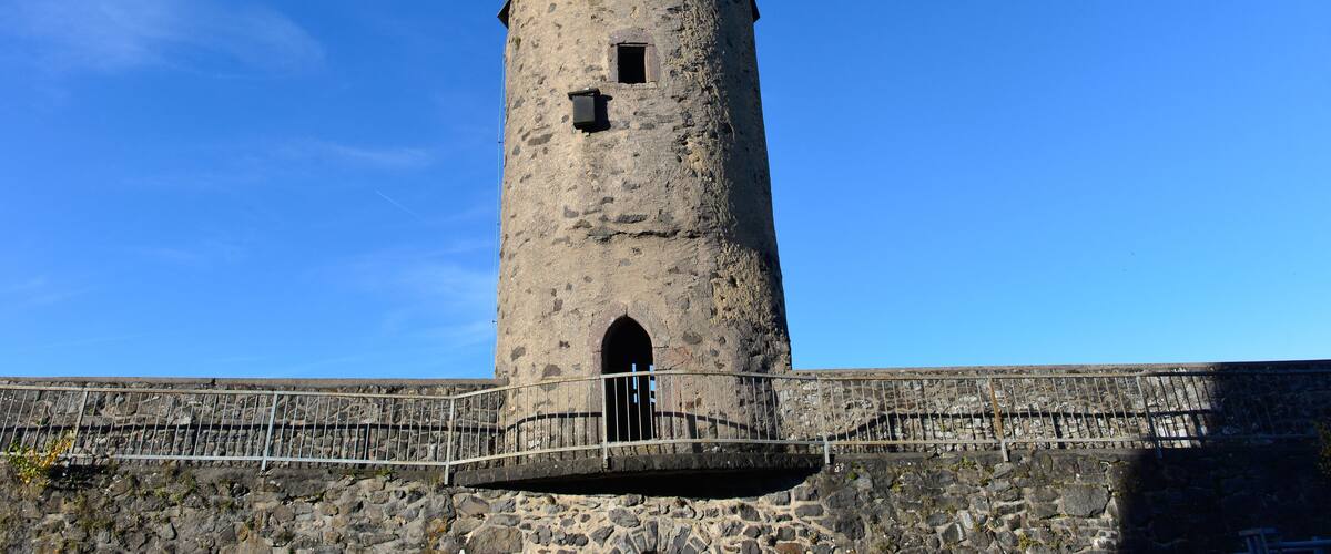 Una alta torre de piedra cilíndrica con techo cónico, una pequeña ventana y una puerta, rodeada por un muro de piedra y una barandilla metálica.