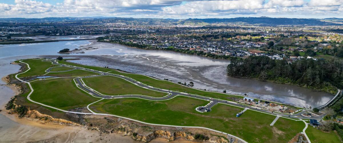 An aerial view of a coastal area with a river, green park, and a city in the background.