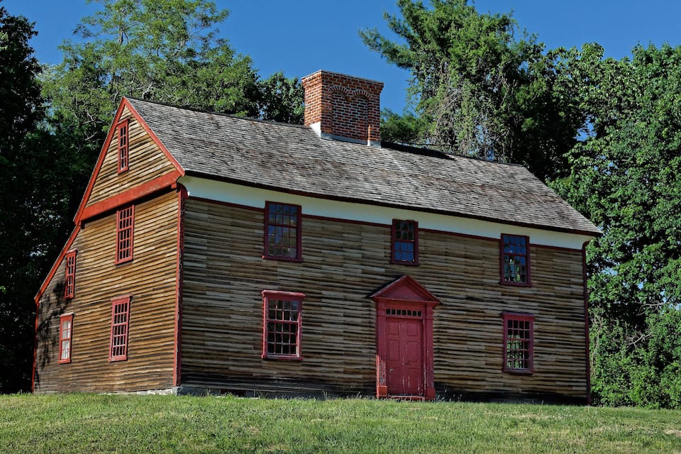A wooden log cabin with a red door and windows, surrounded by green grass and trees.
