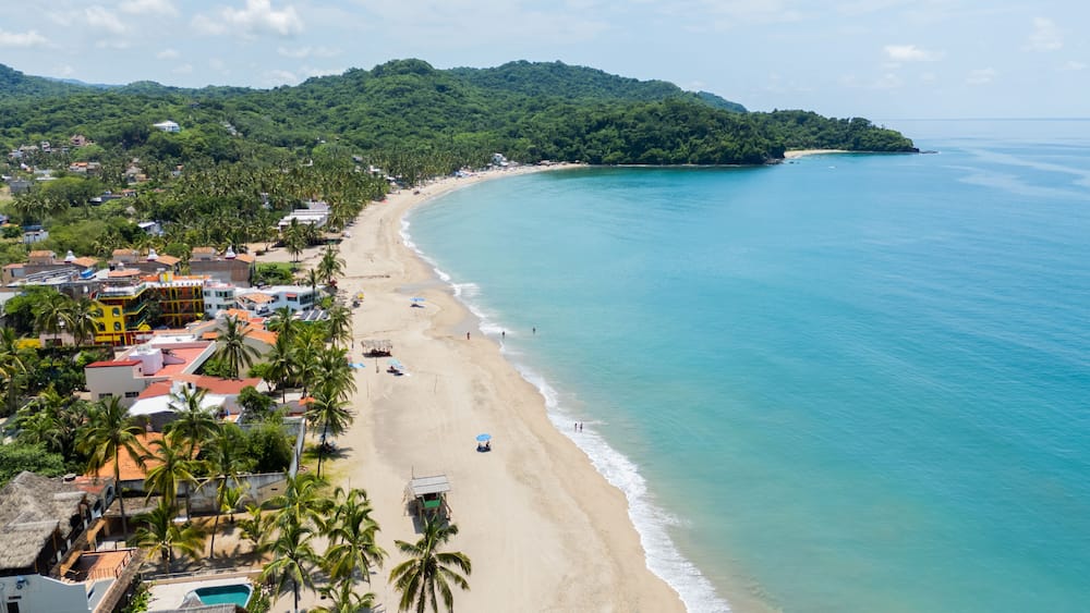 A coastal town with a sandy beach, palm trees, and a mountainous backdrop.