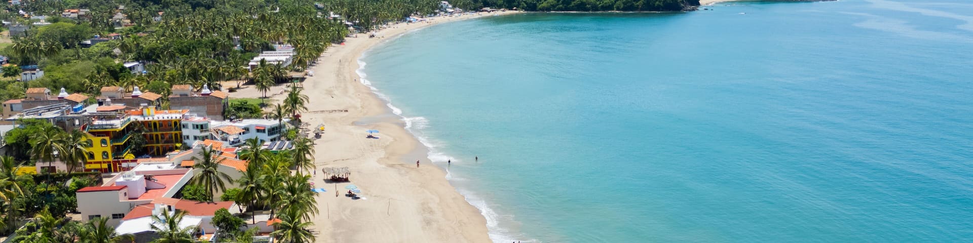 A coastal town with a sandy beach, palm trees, and a mountainous backdrop.
