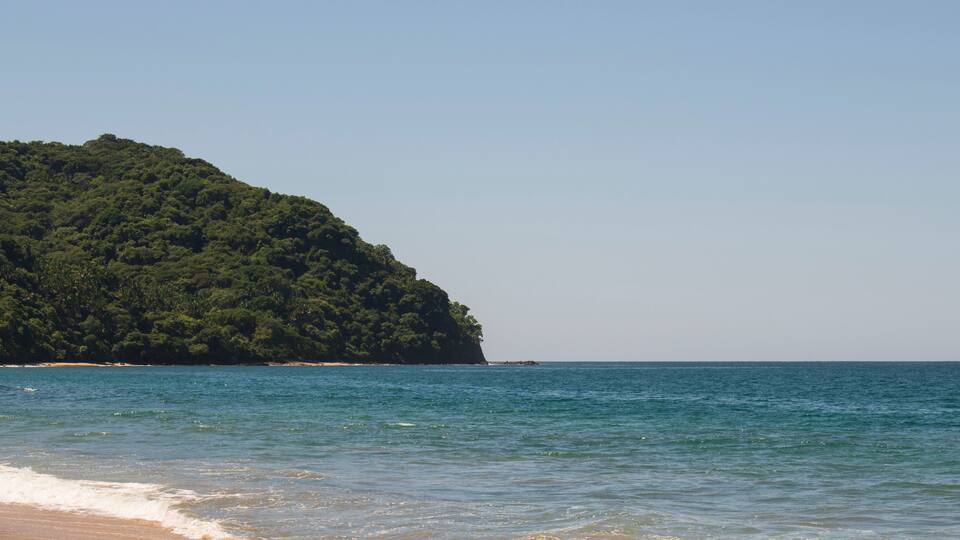 Beautiful landscape of a beach in the mexican pacific coast in a sunny day with a blue and clear sky, trees, palms and green hills, some waves and sand in the beach