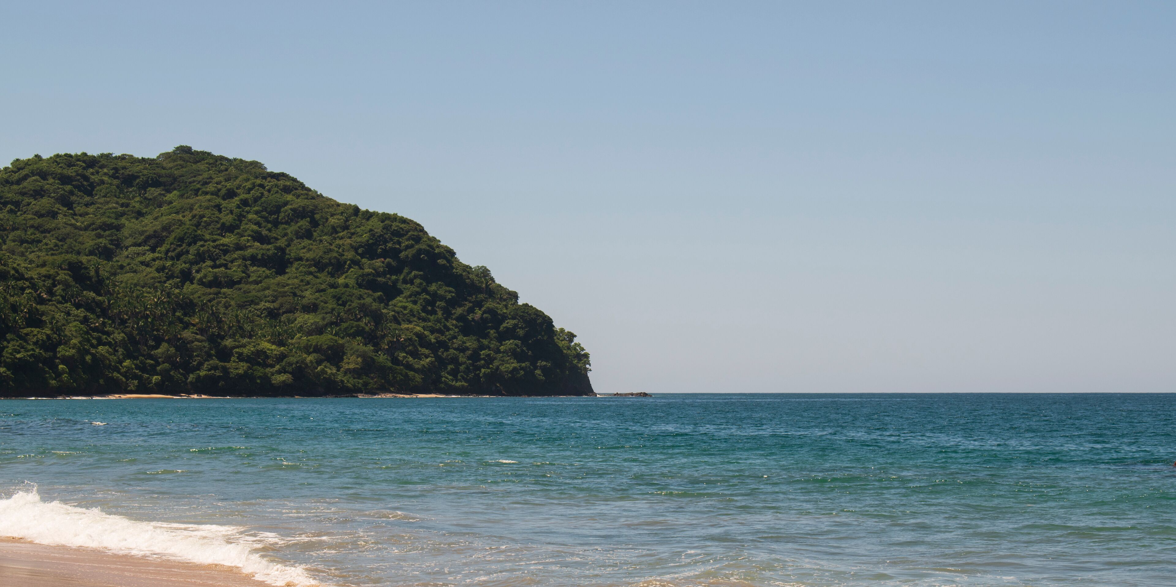 A beach with clear blue water and a large green hill in the background.