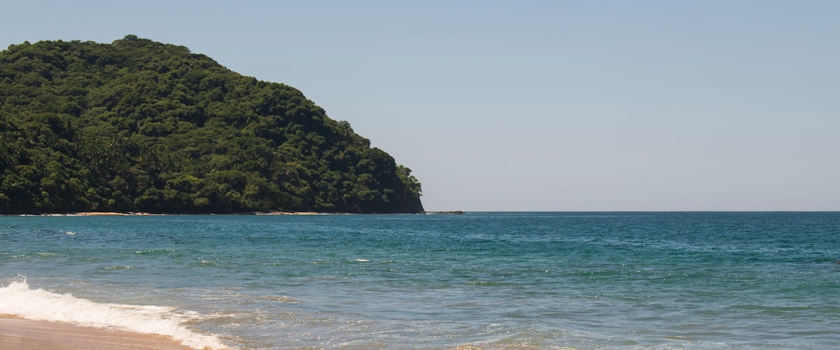 A beach with clear blue water and a large green hill in the background.