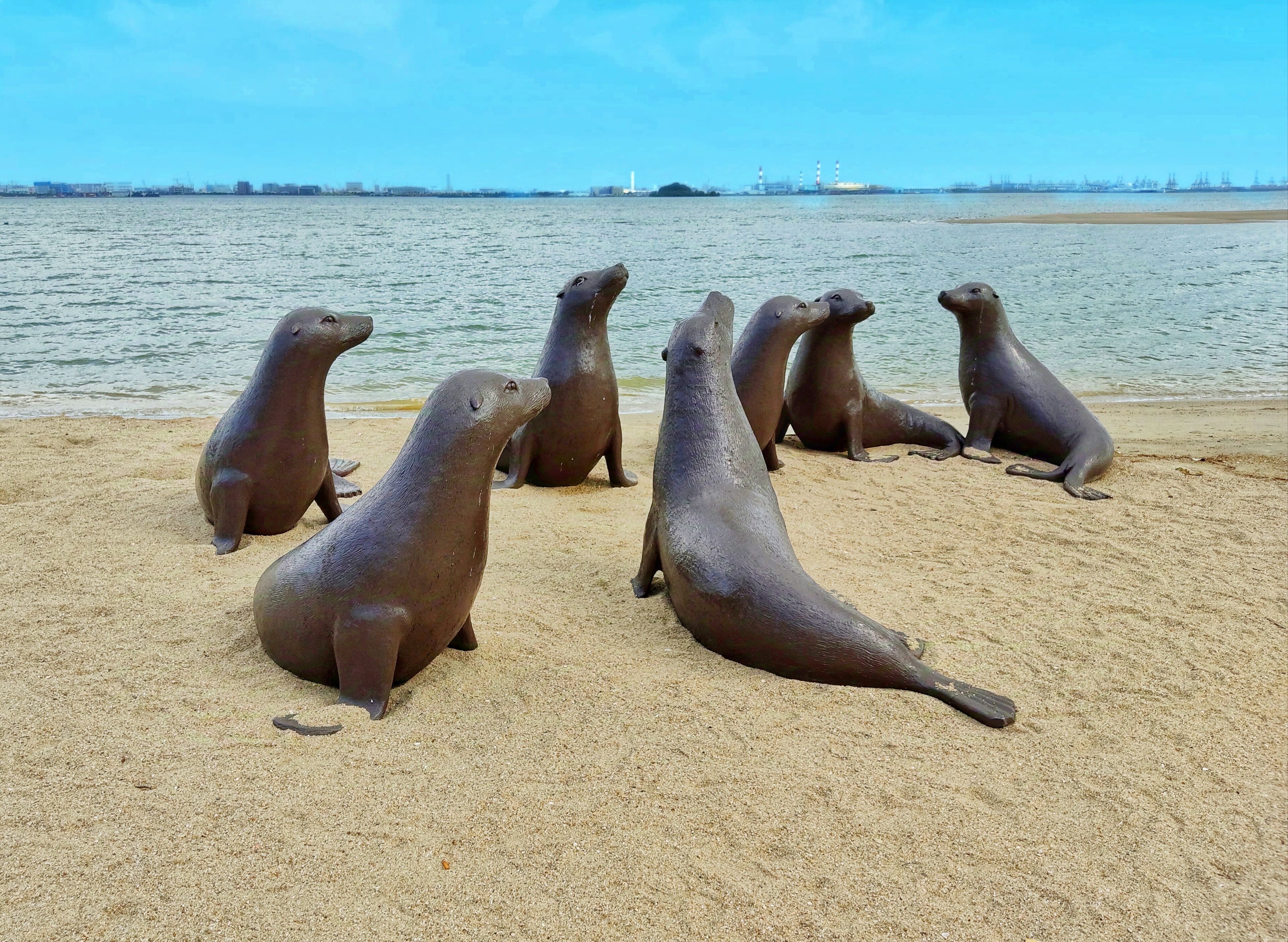 A group of sea lions on a sandy beach with a calm sea and industrial structures in the background.