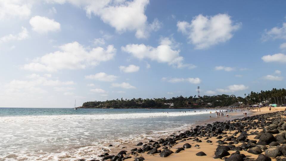 A beach with rocky shoreline, palm trees, and a distant lighthouse.