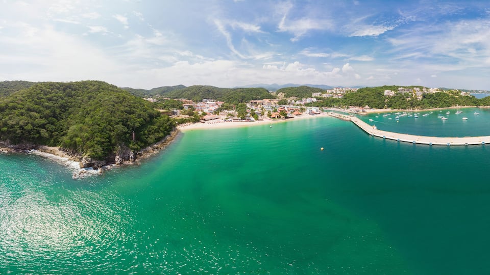 A coastal landscape with a small island surrounded by turquoise waters, a pier extending into the sea, and a backdrop of hills with buildings.