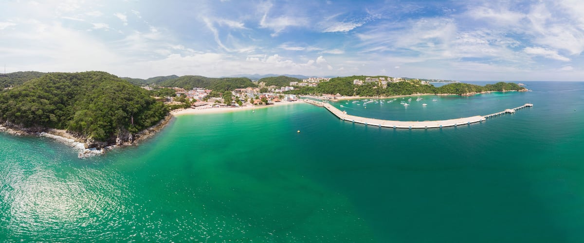 A coastal landscape with a small island surrounded by turquoise waters, a pier extending into the sea, and a backdrop of hills with buildings.