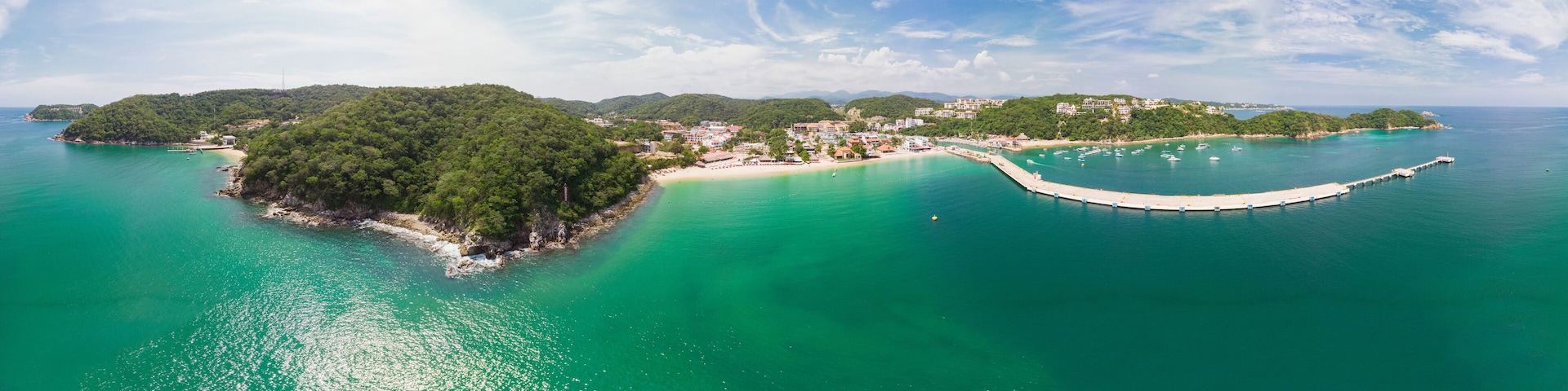 A coastal landscape with a small island surrounded by turquoise waters, a pier extending into the sea, and a backdrop of hills with buildings.