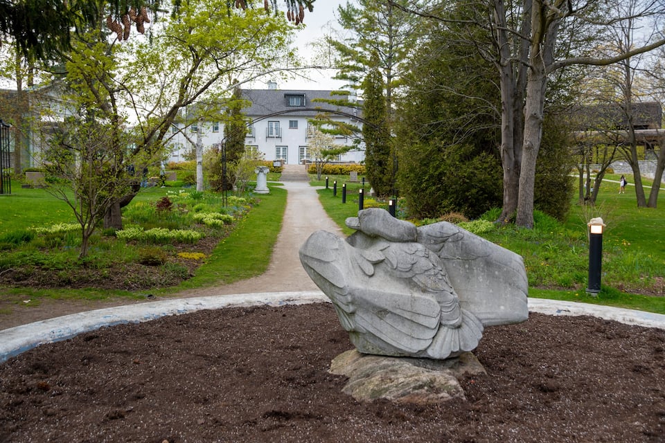 Ein Garten mit einem Weg, der zu einem weißen Gebäude führt. Im Vordergrund steht eine Steinskulptur.
