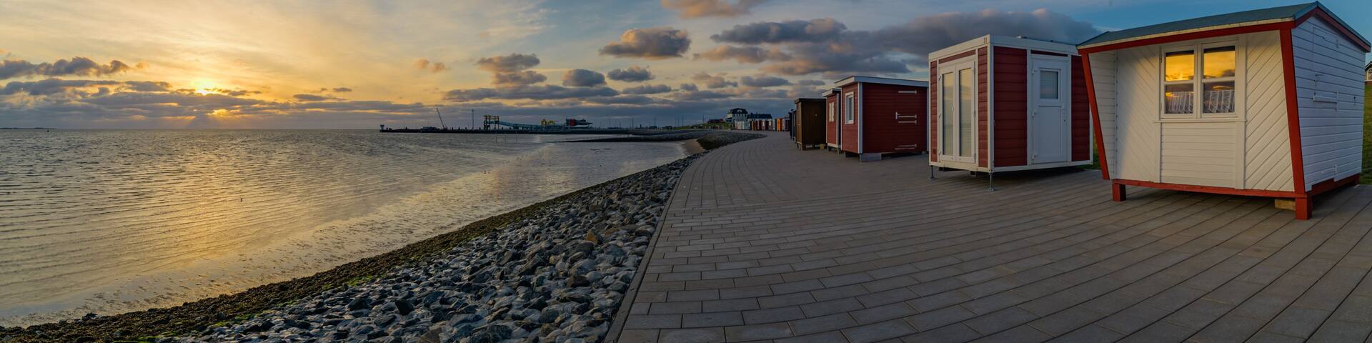 Panorama view of colorful beach houses and bathhouses on the dike of Dagebüll and its harbour on the background. Sunset reflection in the window
of beach house on the dike at the North Sea.