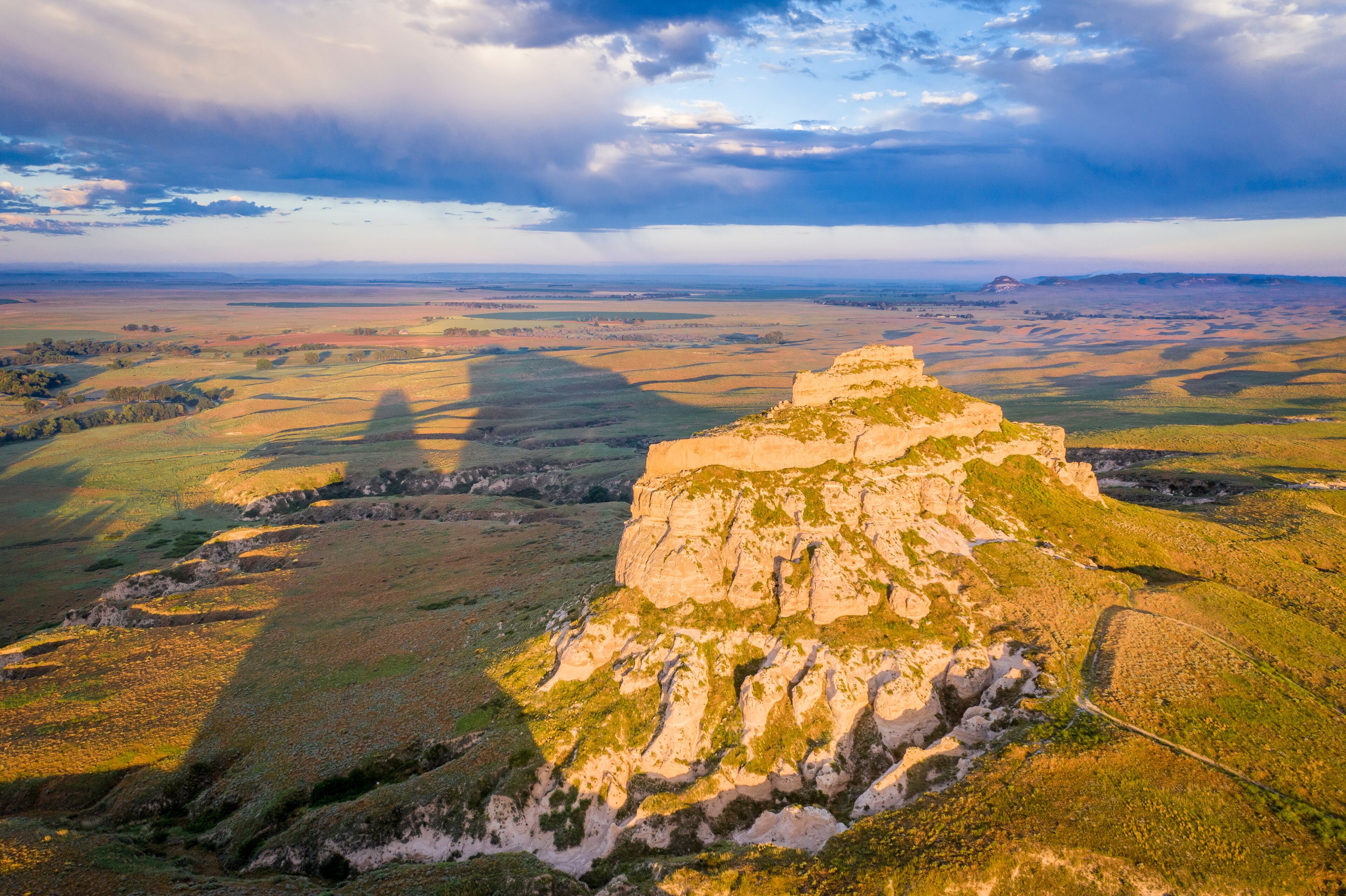 A rugged mountain peak with a winding road leading up to it, surrounded by rolling hills and a vast open landscape under a cloudy sky.