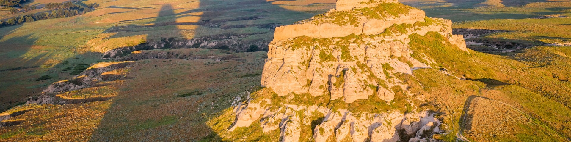 A rugged mountain peak with a winding road leading up to it, surrounded by rolling hills and a vast open landscape under a cloudy sky.